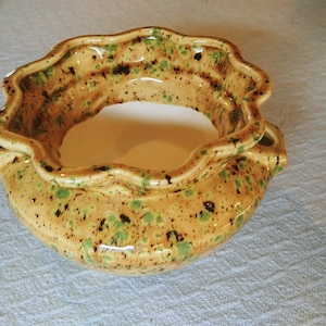 May include: A handcrafted ceramic bowl with a scalloped rim. The bowl has a speckled tan glaze with green and brown accents. The interior is a creamy white. The bowl has two small handles on either side.