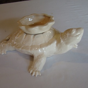 May include: Cream-colored ceramic turtle-shaped decorative bowl. The turtle has a textured shell and detailed legs and head. A smaller, scalloped-edged bowl sits on the turtle's back. The piece is glazed and has a glossy finish.