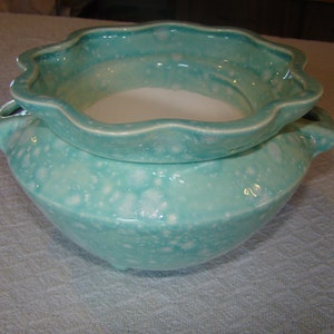 May include: A two-piece ceramic bowl set in a speckled turquoise glaze. The bottom bowl has a rounded shape with small handles, while the top bowl has a scalloped rim. The set is designed for serving or display.