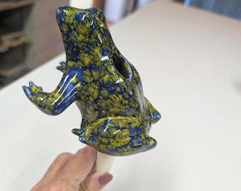 May include: A ceramic frog figurine with a glossy finish featuring a marbled pattern of deep blue and olive green. The frog is sculpted in a seated position with its front legs extended and is attached to a light-colored cylindrical base.