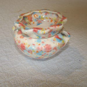 May include: A small, ceramic trinket dish with a lid. The dish and lid are white with colorful speckled patterns in red, blue, and orange. The dish has two small handles.