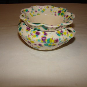 May include: A two-piece ceramic bowl set. The bowls are off-white with a speckled pattern of teal, purple, and yellow dots. The top bowl has a scalloped rim. The bottom bowl has two small handles.