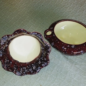May include: Two ceramic bowls with a speckled maroon exterior and a cream-colored interior. One bowl has a scalloped edge, while the other has two small handles. The bowls are likely for serving or display.