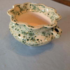 May include: A ceramic wax warmer with a scalloped rim and a cream-colored base featuring green and yellow speckles. The bowl has a wide opening, and a small circular hole is visible on the side of the rounded body for heat ventilation.