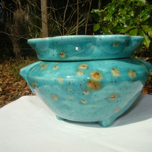 May include: A turquoise ceramic bowl with a matching lid. The bowl has a speckled pattern with brown accents and sits on three small feet. The lid has a scalloped edge. The bowl is likely used for decorative or functional purposes.