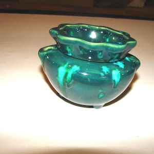 May include: A two-piece, dark green ceramic container with a glossy finish. The top piece is smaller and sits atop a larger, rounded base. Teal accents drip down the sides, adding visual interest. The container has a scalloped edge.