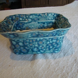May include: A rectangular ceramic dish with a unique, textured surface. The dish is primarily turquoise with a speckled pattern of darker blue and brown. The rim has a wavy design. This decorative piece could be used for serving or display.