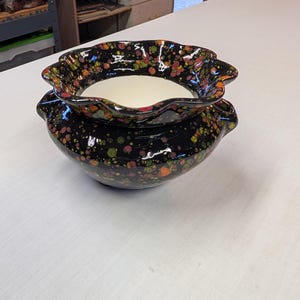 May include: A two-piece ceramic planter set. The bottom piece is a round, black bowl with small handles. The top piece is a ruffled-edge bowl that sits inside the bottom bowl. Both pieces are decorated with colorful speckles.