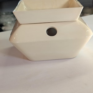 May include: A two-piece, off-white ceramic wax warmer. The base is hexagonal with a circular opening. A rectangular dish sits on top. The warmer is designed to melt wax for fragrance.