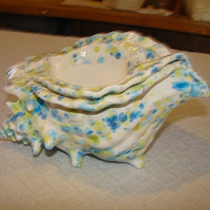 May include: A ceramic seashell-shaped container with a white base, speckled with blue and yellow paint. The container has a textured surface and is supported by three small feet. The interior is a smooth, white bowl.
