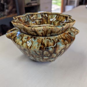 May include: A ceramic bowl with a scalloped rim and two small side handles, featuring a glossy glaze with a marbled pattern of brown, tan, and light blue swirls. The bowl has a rounded body and sits on small feet, showcasing a smooth finish.