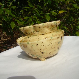 May include: A speckled ceramic bowl with a lid. The bowl is light yellow with brown and green speckles. The bowl has three small feet and a slightly curved rim. The lid is a smaller version of the bowl, with the same color and speckle pattern.