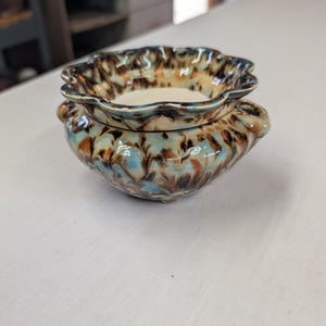 May include: A small ceramic bowl with a scalloped rim and two side handles. The exterior features a mottled glaze in shades of light blue, brown, and cream, while the interior base is a smooth, solid cream color with a glossy finish.