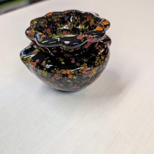 May include: A small, black ceramic pot with a speckled floral pattern in shades of red, orange, and green. The pot has a unique, scalloped rim and a rounded base. The glossy finish enhances the vibrant colors.