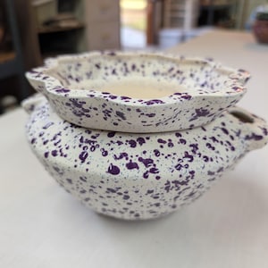 May include: A white ceramic bowl and lid set with a speckled purple pattern. The bowl has two handles and a scalloped rim. The lid has a similar scalloped edge and fits snugly on top of the bowl. The set is likely handmade.