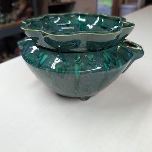May include: A two-piece ceramic planter with a deep emerald green glaze and a glossy finish. The top section features a scalloped rim, while the base bowl has a rounded body with small feet and side handles for easy lifting and handling.