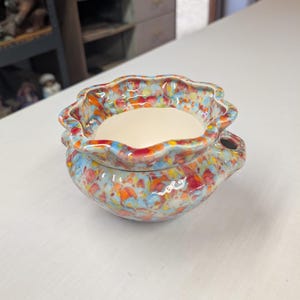 May include: A small ceramic bowl with a scalloped rim and two side handles. The exterior features a vibrant, multi-colored speckled glaze in shades of blue, orange, red, and yellow, while the interior has a smooth, solid cream-colored finish.
