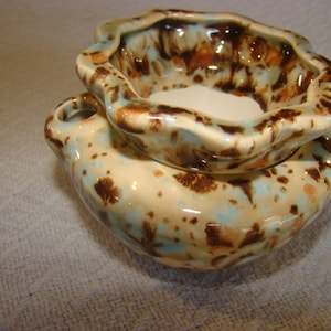 May include: A ceramic decorative bowl with a speckled design. The bowl has a cream-colored base with brown, blue, and orange speckles. The top rim has a wavy design. The bowl has two small handles on the sides.