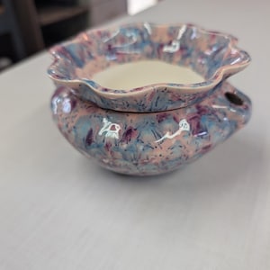 May include: A ceramic wax warmer featuring a scalloped rim and a mottled glaze in shades of pink, blue, and purple. The bowl has a smooth, cream-colored interior basin, while the exterior body displays a textured, multi-colored speckled finish.