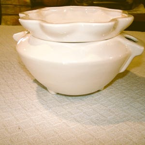 May include: A white ceramic oil warmer with a two-piece design. The top piece has a scalloped edge, and the bottom piece has a rounded shape with three small feet. The warmer has two small handles.