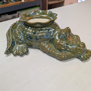May include: A ceramic alligator-shaped oil warmer in shades of green and blue. The alligator has a textured, scaly body and a round dish on its back. The dish is designed to hold oil or wax for aromatherapy.