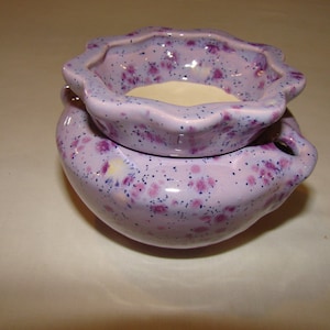 May include: A two-piece ceramic oil warmer in a soft lavender hue, speckled with dark purple and blue dots. The top dish has a scalloped edge and sits atop a rounded base with small handles. The interior of the top dish is a creamy white.
