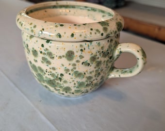 May include: A ceramic mug with a rounded body and a thick, flared rim. The exterior features a cream-colored glaze decorated with a speckled pattern of dark green, light green, and small yellow dots. It includes a single curved handle on the side.