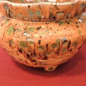 May include: A two-piece ceramic container with a lid. The container is orange with green and black speckled patterns. The base has three small feet. The lid has a scalloped edge.