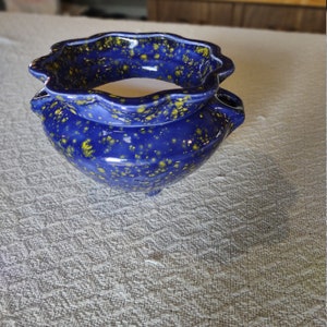 May include: A handcrafted, blue ceramic planter with a speckled yellow pattern. The planter has a wavy rim and three small feet. The interior is white. A small, round opening is on the side of the planter.