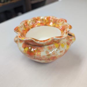 May include: A small ceramic bowl with a ruffled, scalloped rim and a rounded body. The exterior features a mottled glaze pattern in shades of orange, yellow, and white, while the interior is a smooth, solid off-white color.