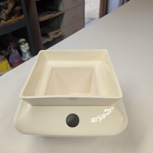 May include: A cream-colored ceramic wax warmer featuring a square-shaped top dish for wax melts and a smooth, rounded base with a single circular opening on the front side for a light source or power cord access, finished in a glossy glaze.