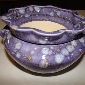 May include: A two-piece ceramic pottery set. The pieces are a deep purple with a speckled pattern of light blue and yellow. The top piece has a scalloped edge. The interior of the top piece is beige.