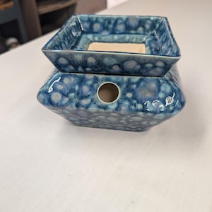 May include: A square ceramic wax warmer featuring a crystalline glaze in shades of blue and white. The object consists of a base with a circular opening on the front side and a removable square dish on top with a smooth, light-colored interior surface.