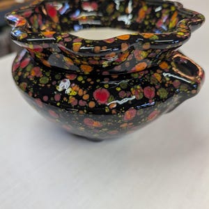 May include: A ceramic vessel with a black base and a speckled pattern of red, orange, yellow, and green dots. The rim has a wavy design. The interior is white. The vessel has a small spout on the side.