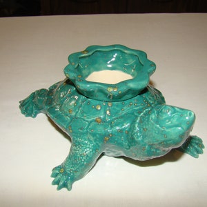 May include: A teal ceramic turtle figurine featuring a textured shell and a scalloped, bowl-shaped top. The glaze is a glossy teal with small, scattered gold-colored speckles throughout the surface, and the interior of the bowl is a smooth, off-white.