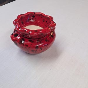 May include: A red ceramic pot with a speckled design. The pot has a wavy rim and a small opening on the side. The interior is white. This decorative piece could be used as a small planter or a unique container for various items.
