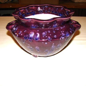 May include: A deep purple ceramic bowl with a ruffled rim and a matching, smaller upper bowl. The glaze has a speckled, glossy finish with blue accents. The bowl sits on a white surface.