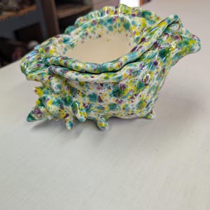 May include: A ceramic seashell-shaped container with a lid. The container is speckled with teal, yellow, and purple paint. The interior is white. The container has a textured surface and is sitting on a white surface.