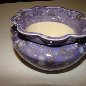 May include: A two-piece ceramic pottery set in shades of purple with yellow speckles. The top piece is a shallow bowl with a scalloped edge, resting on a larger, rounded base. The interior of the top bowl is a creamy white color.