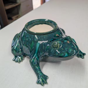 May include: A ceramic frog-shaped planter with a glossy, deep teal and forest green mottled glaze. The frog is depicted in a crouching position with a circular opening on its back. The interior of the planter is a smooth, off-white ceramic finish.
