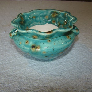 May include: A turquoise ceramic bowl with a ruffled rim and a speckled, textured surface. The bowl has a white interior and small handles on either side. The bowl is likely handmade and suitable for decorative or functional use.