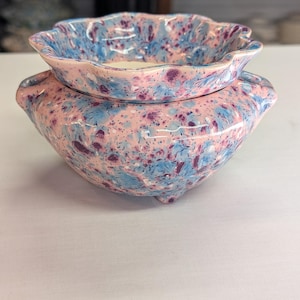 May include: A two-piece ceramic set with a pink, blue, and purple speckled glaze. The bottom piece is a rounded bowl with handles, and the top piece is a smaller, scalloped-edge bowl. The set is likely for decorative or functional use.
