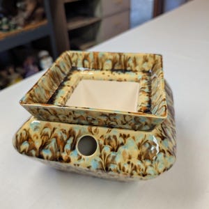 May include: A square ceramic wax warmer featuring a marbled glaze in shades of tan, brown, and light blue. The base has a circular opening on the front, and a removable square dish with a solid white interior sits on top of the main body.