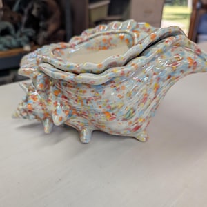 May include: A ceramic seashell-shaped planter with a speckled design. The planter is white with colorful speckles of blue, orange, and red. It has a textured surface and a smooth, white interior. The planter has three small feet.