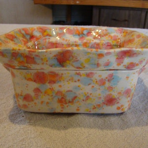 May include: A rectangular ceramic dish with a lid, featuring a speckled pattern of red, blue, orange, and yellow dots on a white background. The dish has a wavy rim and is likely used for serving or storage.