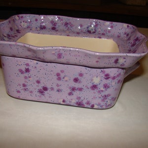 May include: Two-piece ceramic baking dish set. The set features a lavender base with a speckled purple design. The top dish has a scalloped edge and a cream-colored interior. The bottom dish has handles.