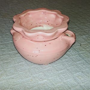 May include: A pink ceramic container with a ruffled top and small handles. The container has a speckled design with dark brown spots. The interior appears to be white. The container is likely used for decorative purposes.