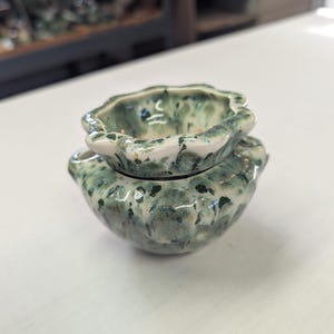 May include: A small, round ceramic oil burner with a scalloped rim and a glossy finish. The piece features a mottled, speckled glaze in shades of sage green, dark forest green, and cream, with a central bowl for oil and a hollow base for a tea light.