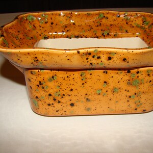 May include: A rectangular ceramic planter with a two-tiered design. The planter is glazed in a speckled orange color with green and black accents. The top tier has a wavy rim, and the interior is white.