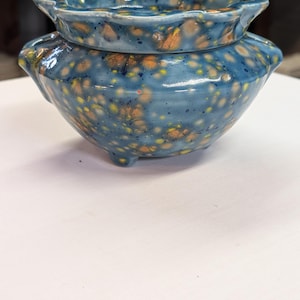 May include: A handcrafted ceramic bowl and lid set. The bowl is a deep blue with a speckled pattern of yellow and orange. The lid has a scalloped edge. The bowl has three small feet.
